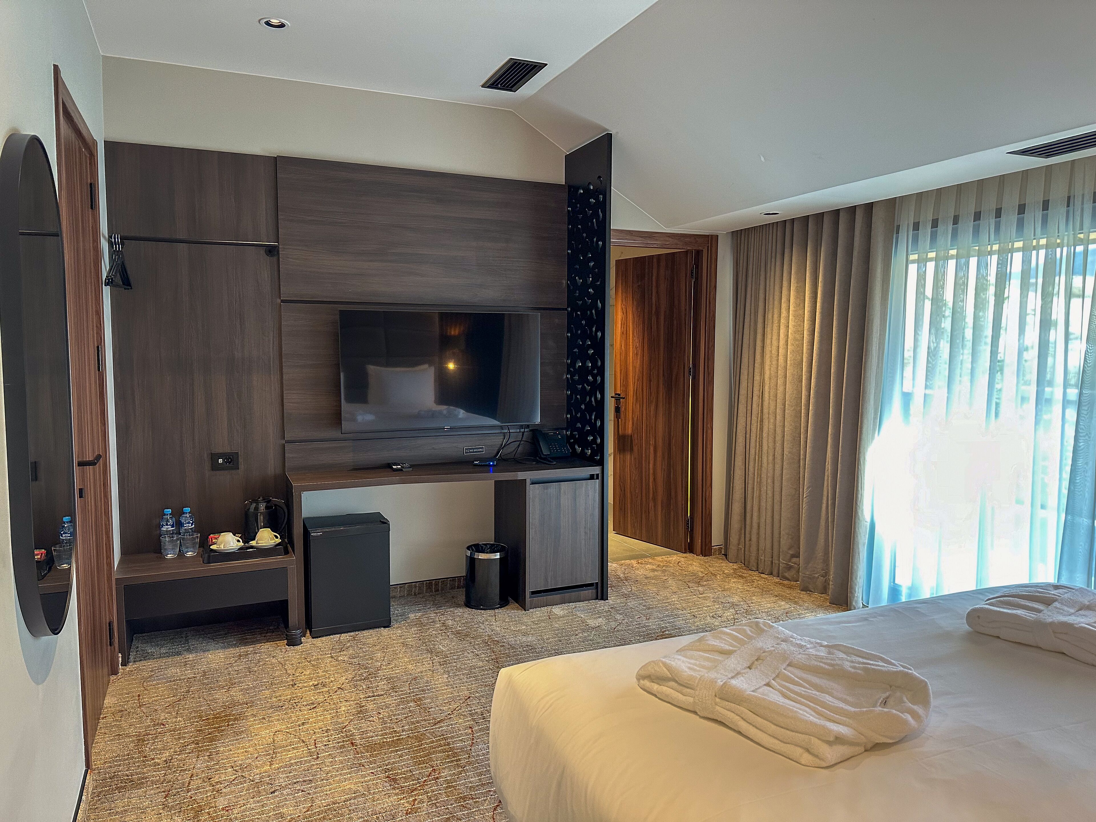 A modern hotel room with a large bed, a flat-screen TV mounted on a wooden panel, a desk with a computer, and a small seating area with a chair and a table.
