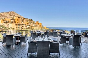 An outdoor dining area with tables and chairs, overlooking a coastal landscape with buildings in the background. - Los Cabos | Couples All-Inclusive Retreat | Pool + Beach (Cabo del Sol)
