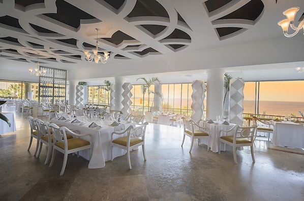 A spacious dining area with white tables and chairs, a modern ceiling design, and a view of the ocean.