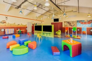 A brightly lit indoor play area with colorful padded seating, tables, and a mural on the wall.