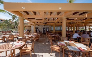 An outdoor dining area with wooden tables and chairs, covered by a pergola with wooden beams and ceiling fans.