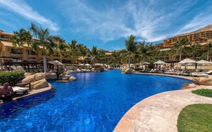 A resort pool area with multiple lounge chairs and umbrellas, surrounded by palm trees and a large building in the background. - Los Cabos | Couples All-Inclusive Retreat | Pool + Beach (Cabo del Sol)