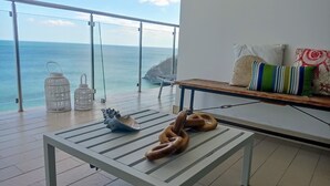 Un balcón con vista al mar, una mesa blanca con una concha decorativa y un banco con cojines.