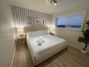 A neatly made bed with a rolled towel, a bedside lamp, a small table with a plant, and a window with blinds. - Surf Shack 2 - 5 min to beach, 12 min to Legoland! (Encinitas)
