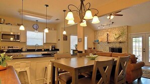 A modern kitchen with a dining area, featuring a stone fireplace, pendant lighting, and a clock on the wall.