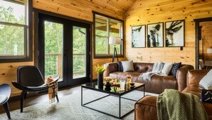 A cozy cabin interior with a brown leather sofa, a glass coffee table, and a large window offering a view of trees. - Fall Mtn Escape! HeatedPool-HotTub-Theater-Views (Sevierville)