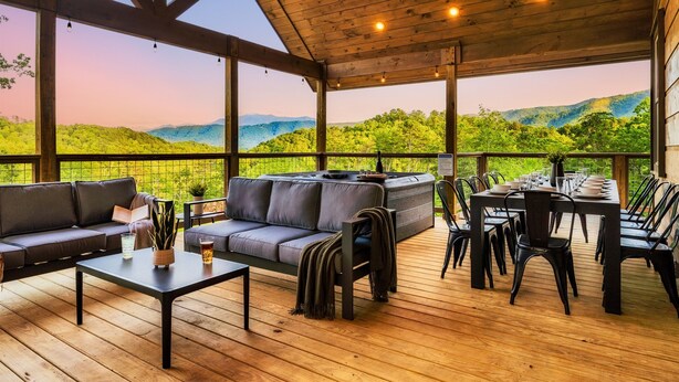 A covered outdoor seating area with a wooden deck, a sofa, a table, and chairs. - Fall Mtn Escape! HeatedPool-HotTub-Theater-Views (Sevierville)