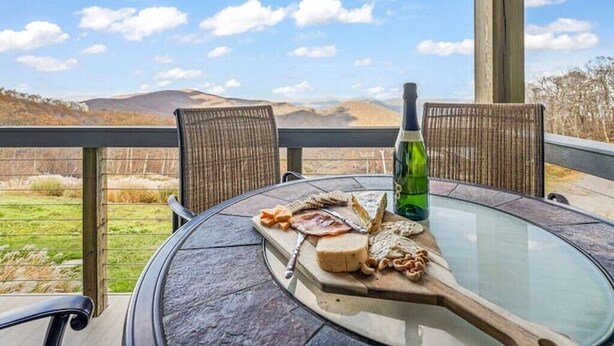Une table ronde en verre, sur laquelle repose une planche en bois présentant du fromage, du pain et une bouteille, est placée sur un balcon agrémenté de chaises en osier et offrant une vue sur des collines.