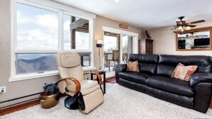 Un salon spacieux doté d’une grande fenêtre, d’un canapé en cuir noir, d’un fauteuil inclinable et d’un ventilateur de plafond.