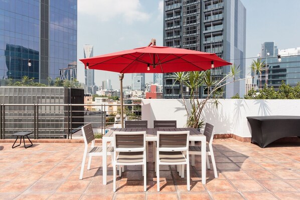 A rooftop terrace with a table and chairs, a red umbrella, and a view of the city skyline.