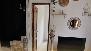 Een kamer met een open haard, een televisie en een deur naar een andere kamer. Er staan decoratieve objecten aan de muur en een plant op een plank.