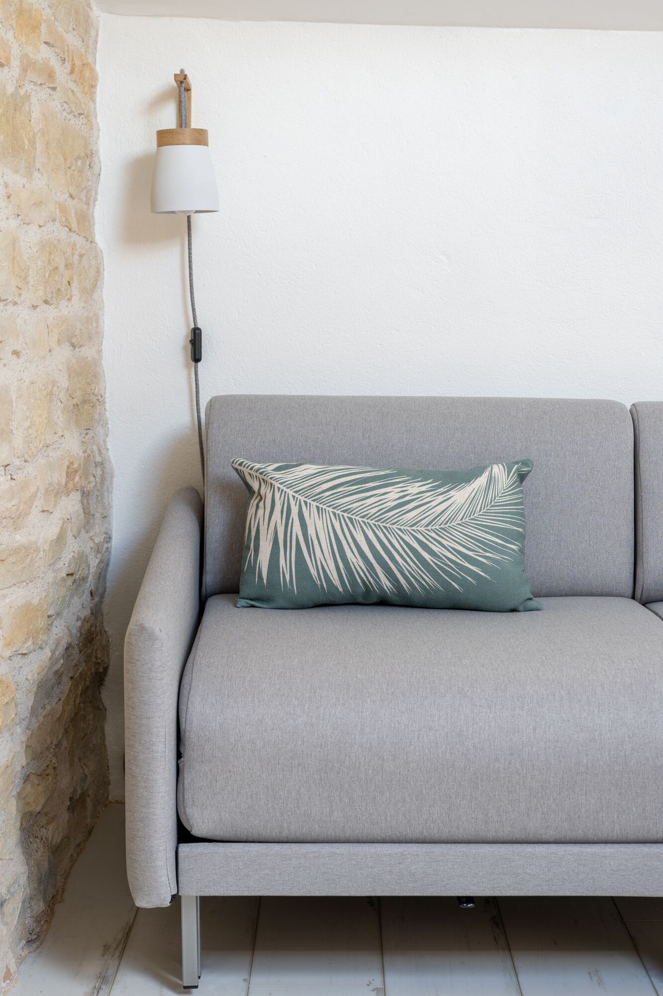 Ein graues Sofa mit einem Kissen aus Palmenblättern, eine weiße Lampe mit Holzfuß und eine Steinwand.