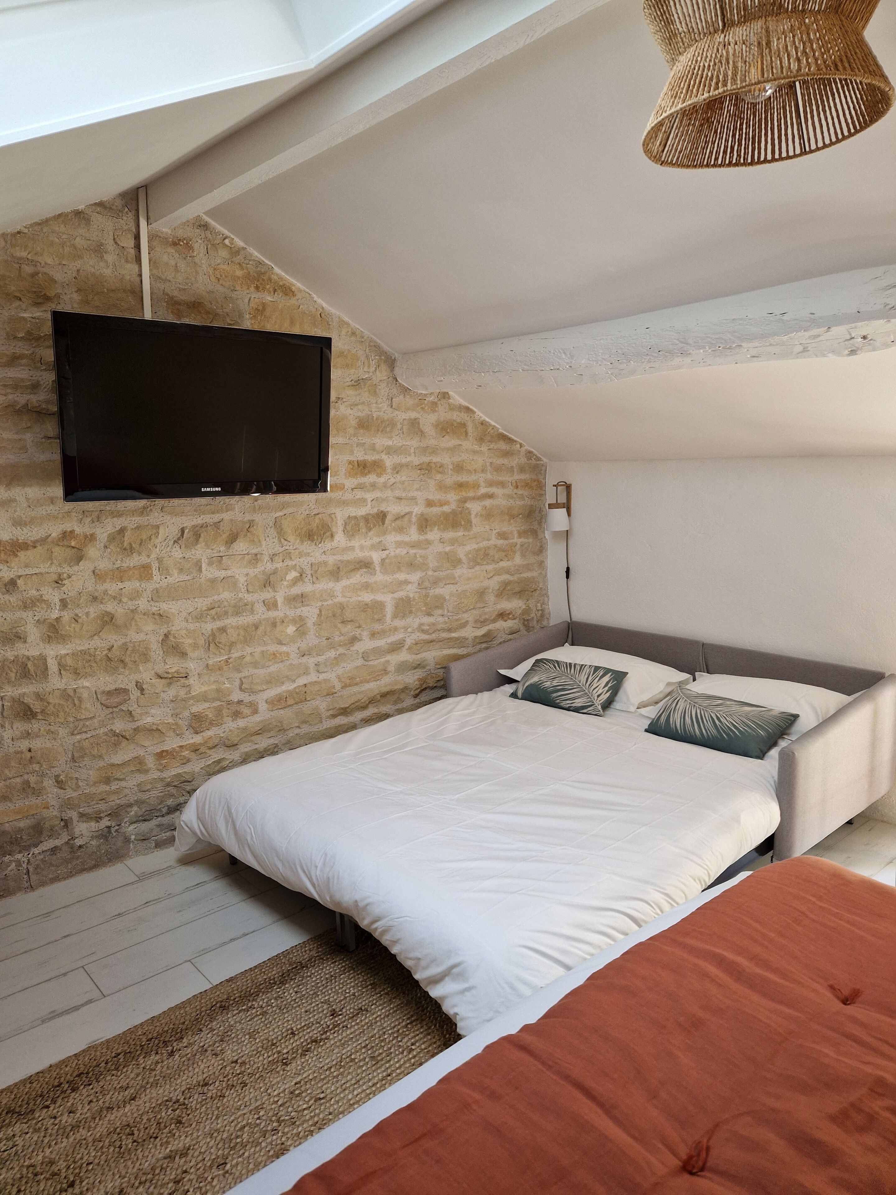 A bedroom with a stone wall, a flat-screen TV mounted above the bed, a woven light fixture, and a bed with white bedding and patterned pillows.