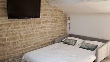 A bedroom with a stone wall, a flat-screen TV mounted above the bed, a woven light fixture, and a bed with white bedding and patterned pillows.
