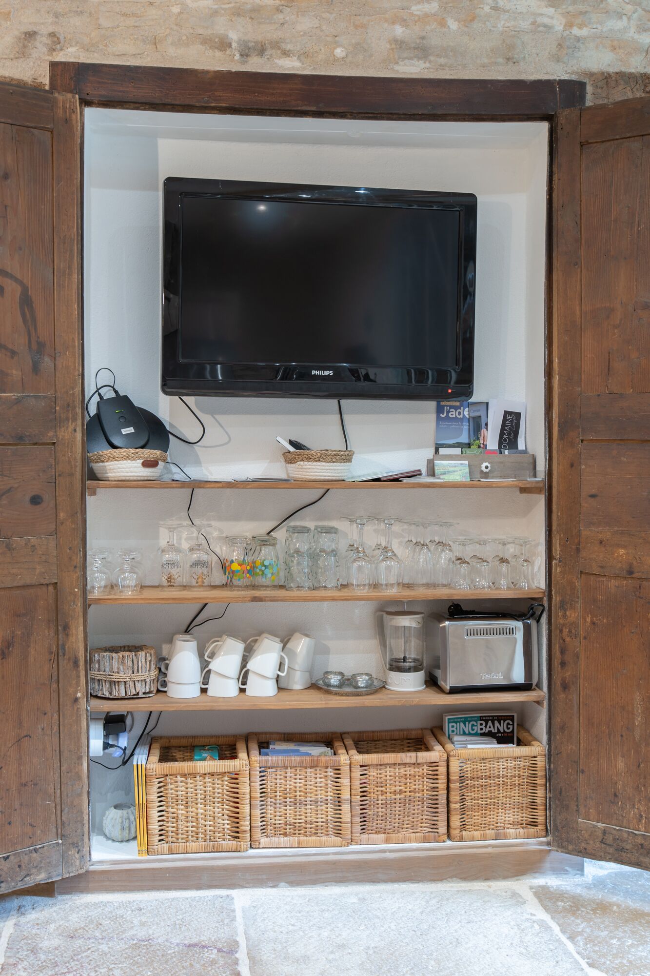 Un téléviseur fixé au mur, avec des étagères en bois en dessous, où sont rangés des verres, des bouteilles et un magazine.