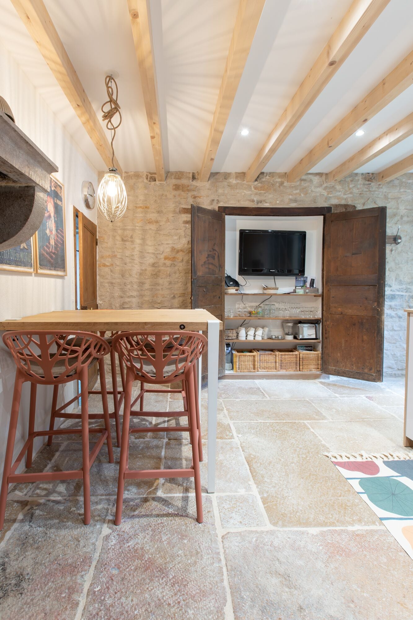 Un intérieur rustique avec des poutres apparentes, un mur en pierre, une table à manger en bois avec des chaises, un téléviseur et des étagères remplies de divers objets.