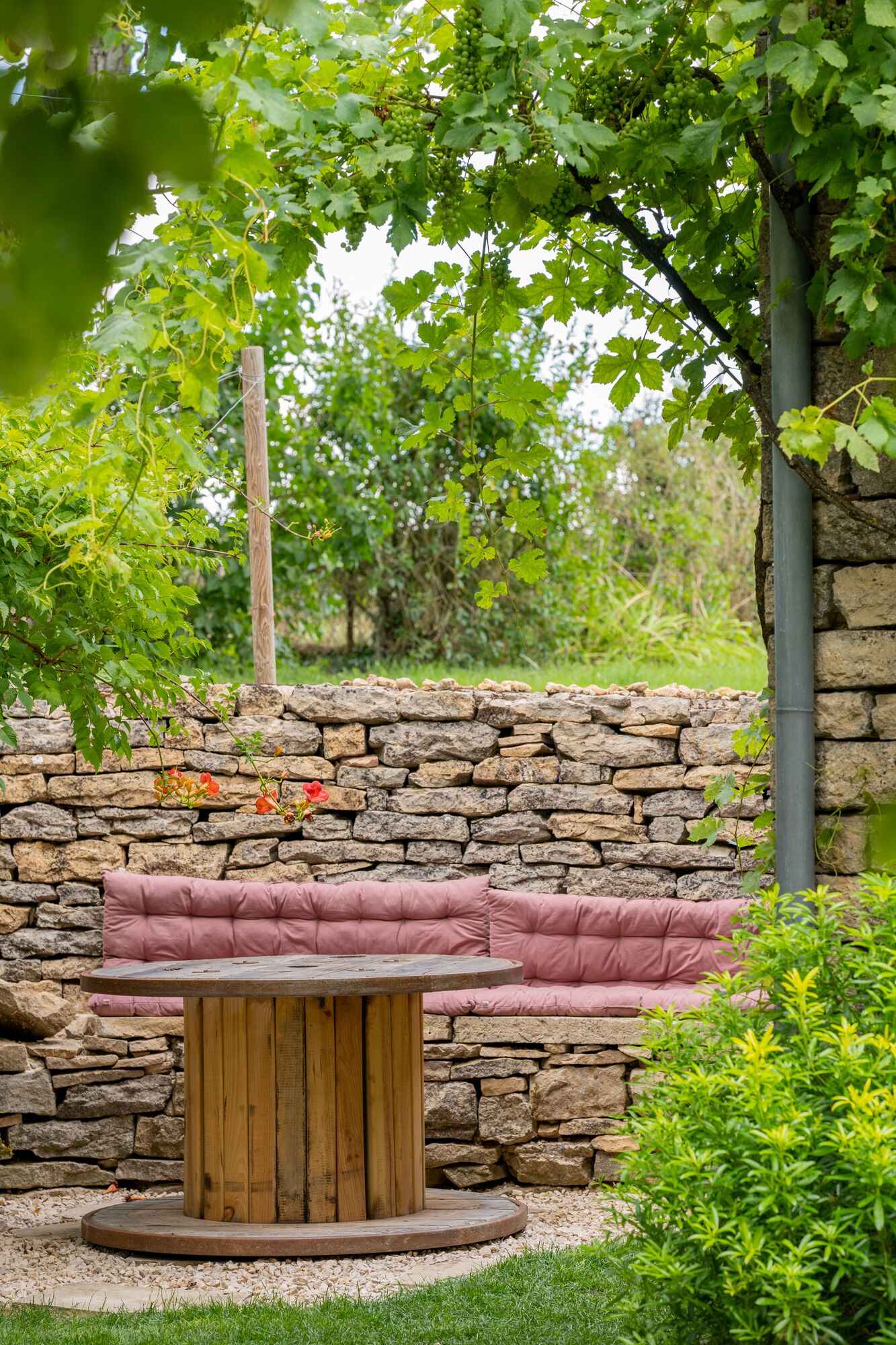 Un mur de pedra, una taula de fusta i una banca amb coixins rosa en un jardí.
