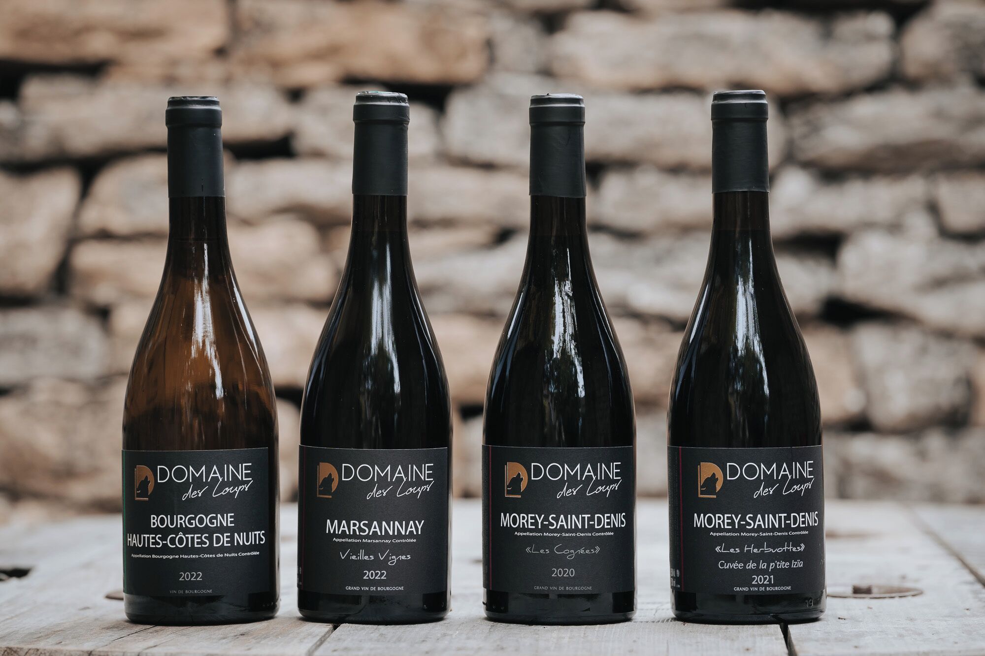 Quatro garrafas de vinho com etiquetas, dispostas sobre uma superfície de madeira, com um muro de pedra ao fundo.