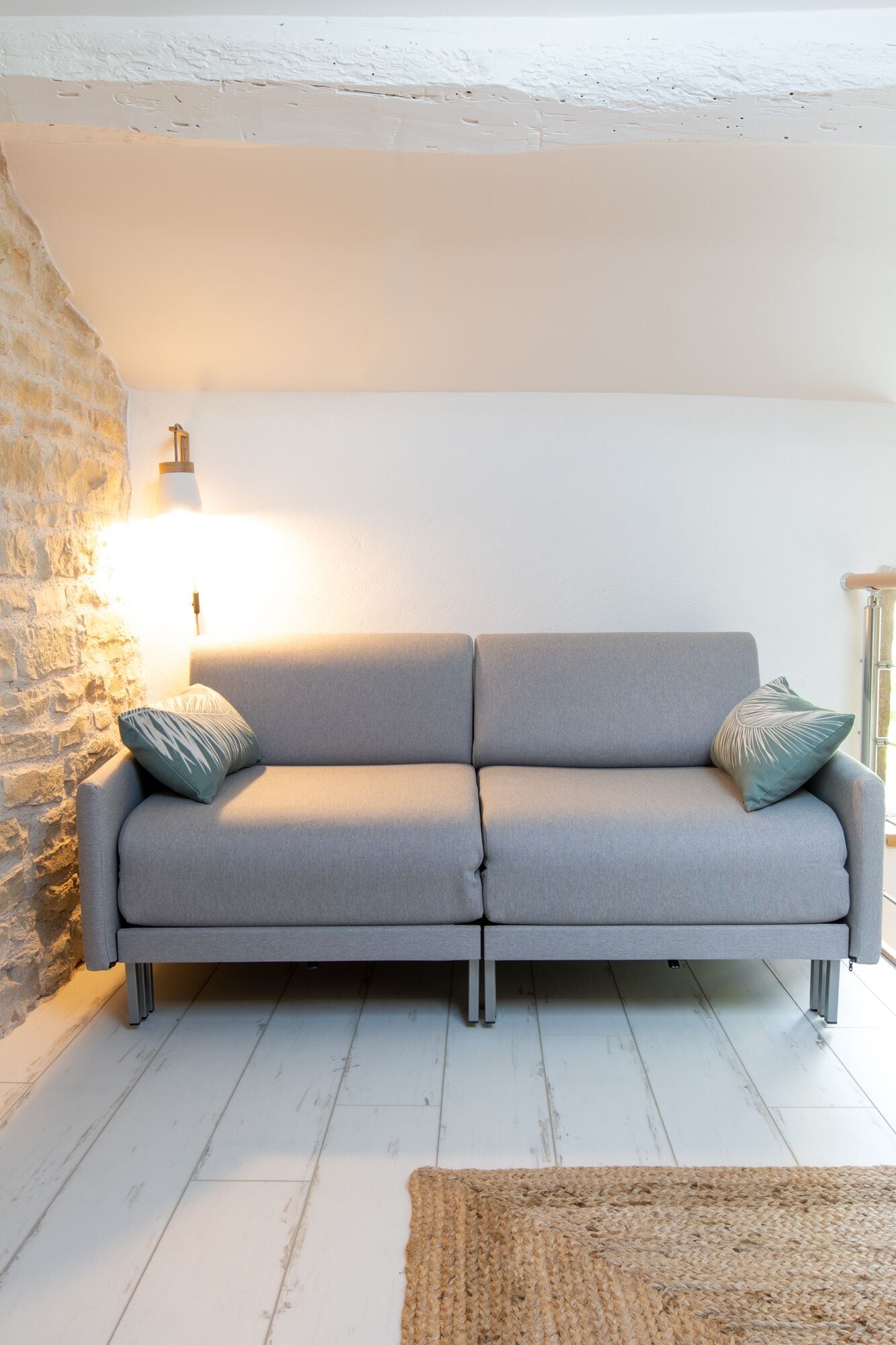 En grå soffa med två kuddar, en vävd matta, en stenmur och en väggmonterad lampa.