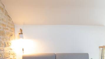 En grå soffa med två kuddar, en vävd matta, en stenmur och en väggmonterad lampa.