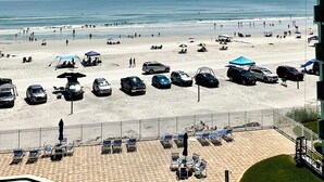 Vista a la playa desde un balcón con terraza pavimentada, sillas de descanso y sombrillas.