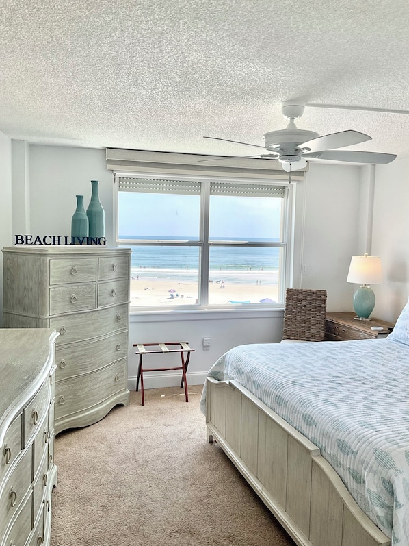 Un dormitorio con vista a la playa, un ventilador de techo, un aparador, una cama y una mesita de noche con una lámpara.
