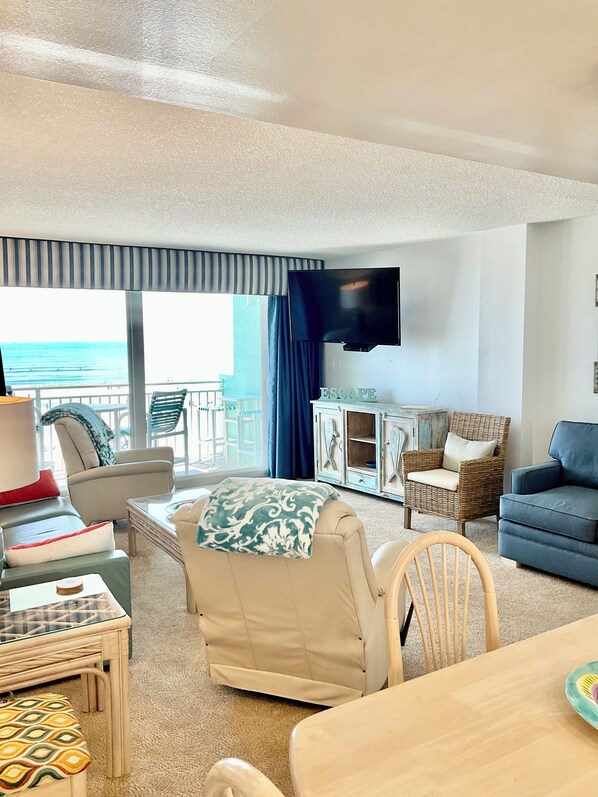 A living room with a view of the ocean, a television mounted on the wall, and a wicker chair.