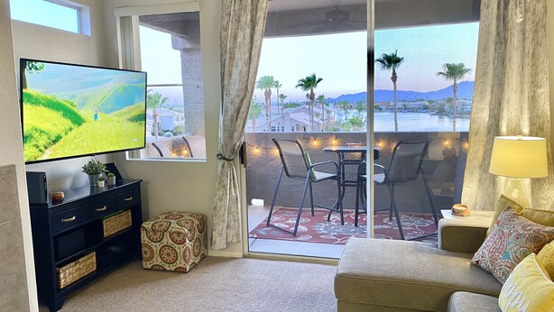 A living room with a TV, a sofa, a dining table with chairs, and a view of a pool and palm trees. - Gorgeous Lakeview Resort Style Condo (Phoenix)