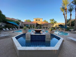 A hotel pool area with a central fountain, lounge chairs, and palm trees.