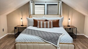 Un dormitorio con una cama, dos mesitas de noche, una lámpara en el techo y una ventana con persianas.