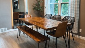 Un comedor con mesa y sillas de madera, un banco, una planta en maceta y una ventana con cortinas.