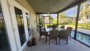 A covered patio with a glass table and wicker chairs, a pool, and a view of the garden.
