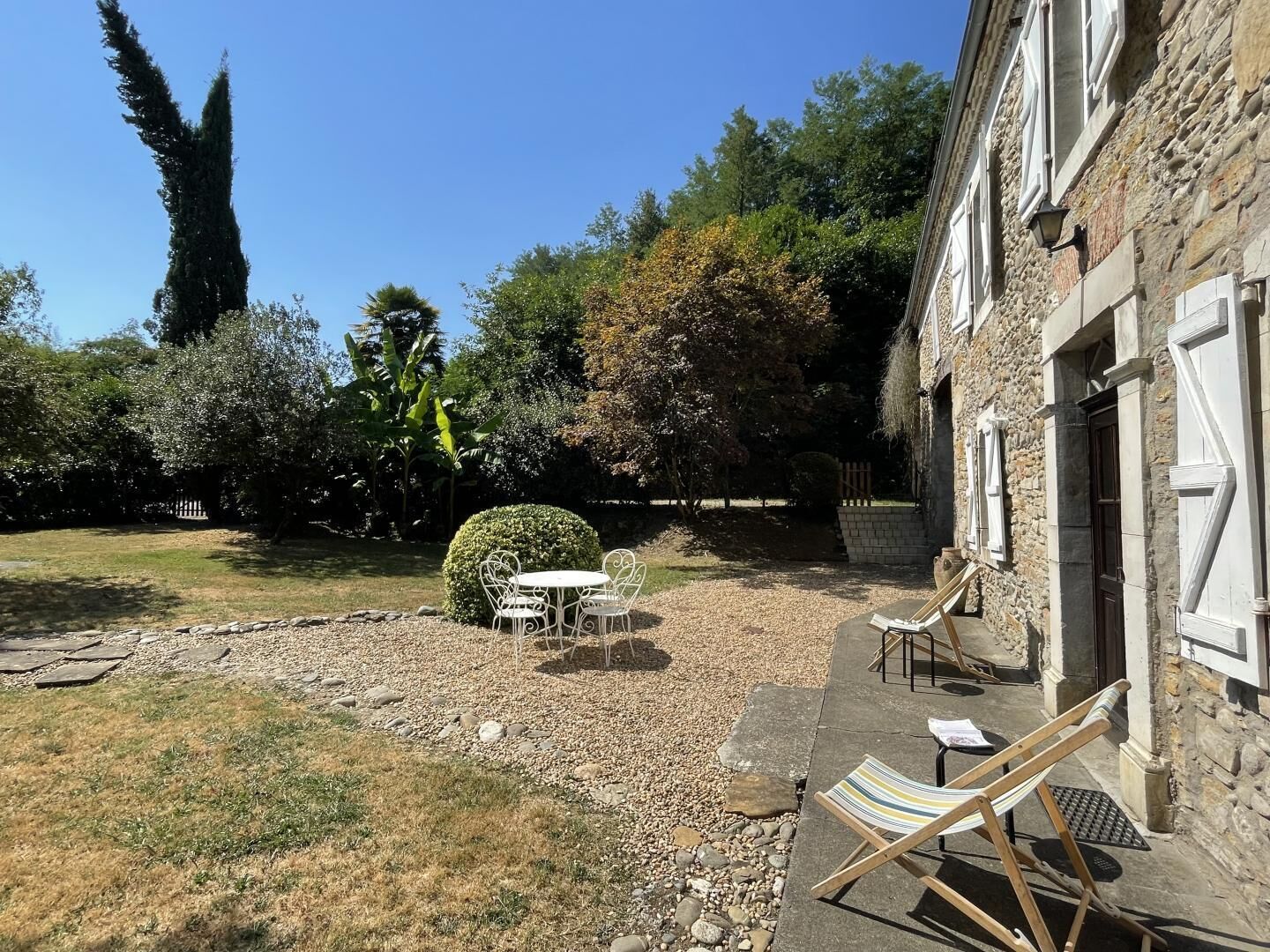 A stone building with a garden patio featuring a table and chairs, surrounded by trees and a hedge.