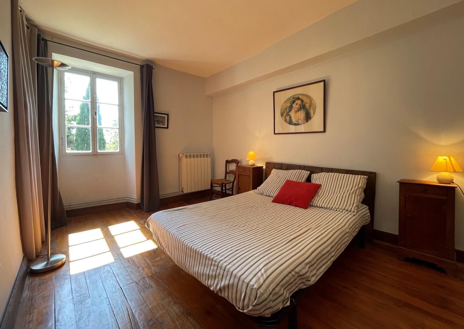 Een slaapkamer met een groot bed, een raam met gordijnen, een radiator, een stoel en twee nachtkastjes met lampen.