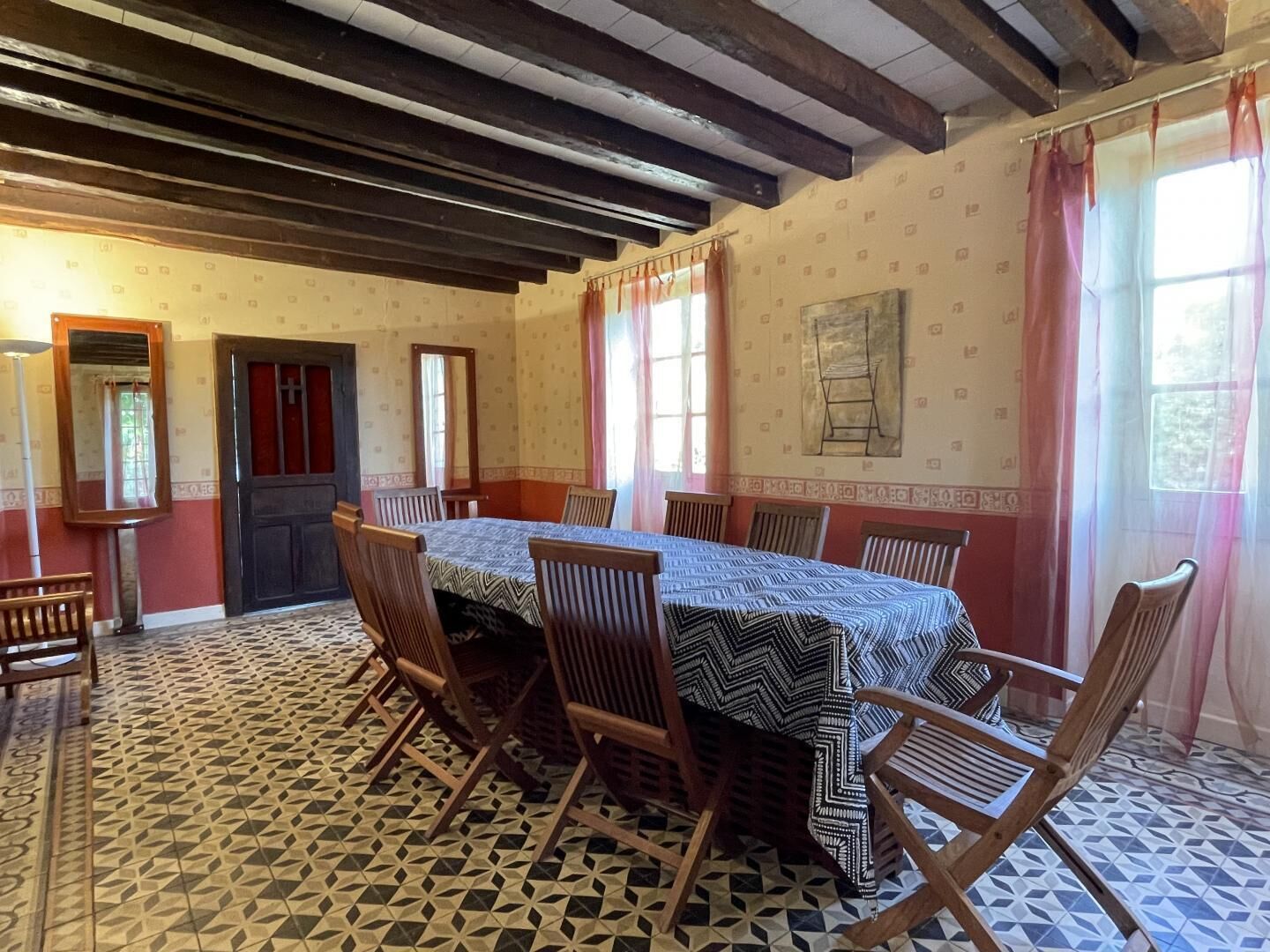 Een eetkamer met een lange tafel, houten stoelen, vloertegels met een patroon en een raam met gordijnen.
