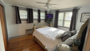 Une chambre à coucher avec un lit, un ventilateur de plafond, un téléviseur fixé au mur et une fenêtre avec des rideaux.