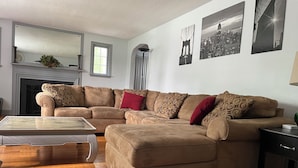 Un salon avec un canapé d’angle beige, une table basse en verre, une cheminée et trois œuvres d’art mural représentant des paysages urbains en noir et blanc.