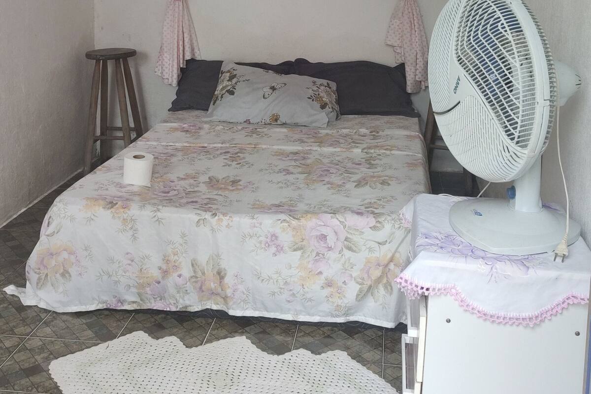A bedroom with a bed, a standing fan, a small stool, a framed picture on the wall, and a doily on the floor.