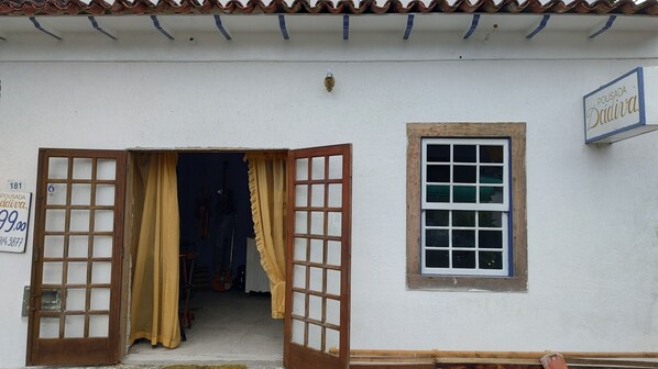A white building with a tiled roof, a wooden door with glass panels, a small window with a white frame, and a sign with text. - Rua São Pedro de Alcântara (Paraty)