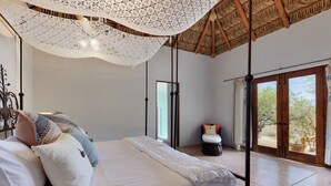 Ein Schlafzimmer mit einem großen Bett, einem Deckenventilator, einem Fenster mit Blick auf Bäume und einer Tür, die in einen anderen Raum führt.
