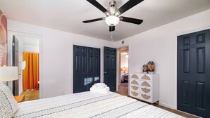 Un dormitorio con cama, ventilador de techo, cómoda blanca y dos puertas oscuras.