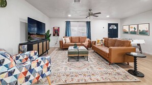 Una sala de estar con un sofá de cuero marrón, un sillón estampado, una mesa de centro y una mesa de fútbol de mesa. Hay un televisor en la pared, un ventilador de techo y una ventana con cortinas.