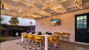 Un patio al aire libre con una mesa blanca dispuesta para seis personas, sillas amarillas, una parrilla y luces decorativas.