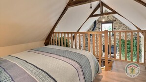 A bedroom with a sloped ceiling, a wooden bed, a view of a stone wall, and a logo on the floor.