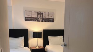 Habitación de hotel con dos camas, una mesita de noche con lámpara, un cuadro enmarcado en la pared y una puerta que da a otra habitación.