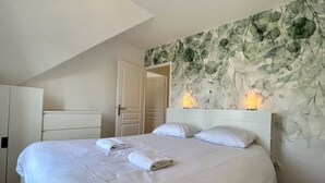 A neatly made bed with white linens and towels, a white headboard, and a wall with a leafy pattern.
