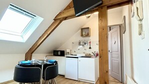 A compact kitchen with a microwave, sink, and white cabinets. A dining table with chairs is positioned under a slanted ceiling with a skylight. - Studio for 2 in the city center (Le Crotoy)