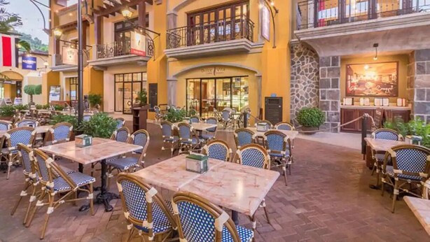 Ein Essbereich im Freien mit mehreren Tischen und Stühlen, einer Steinfassade und einem Restaurant im Hintergrund.