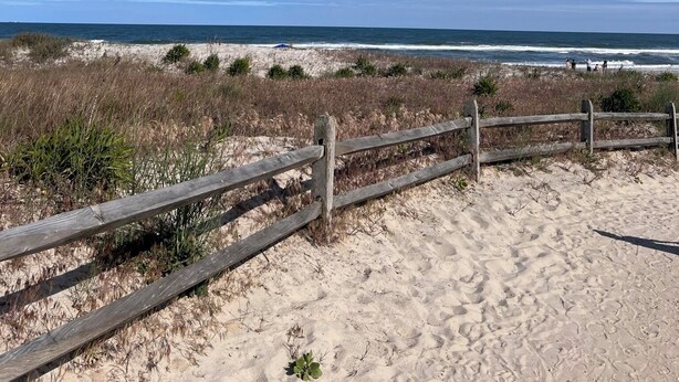 Una playa con una valla de madera, dunas arenosas y el océano al fondo.