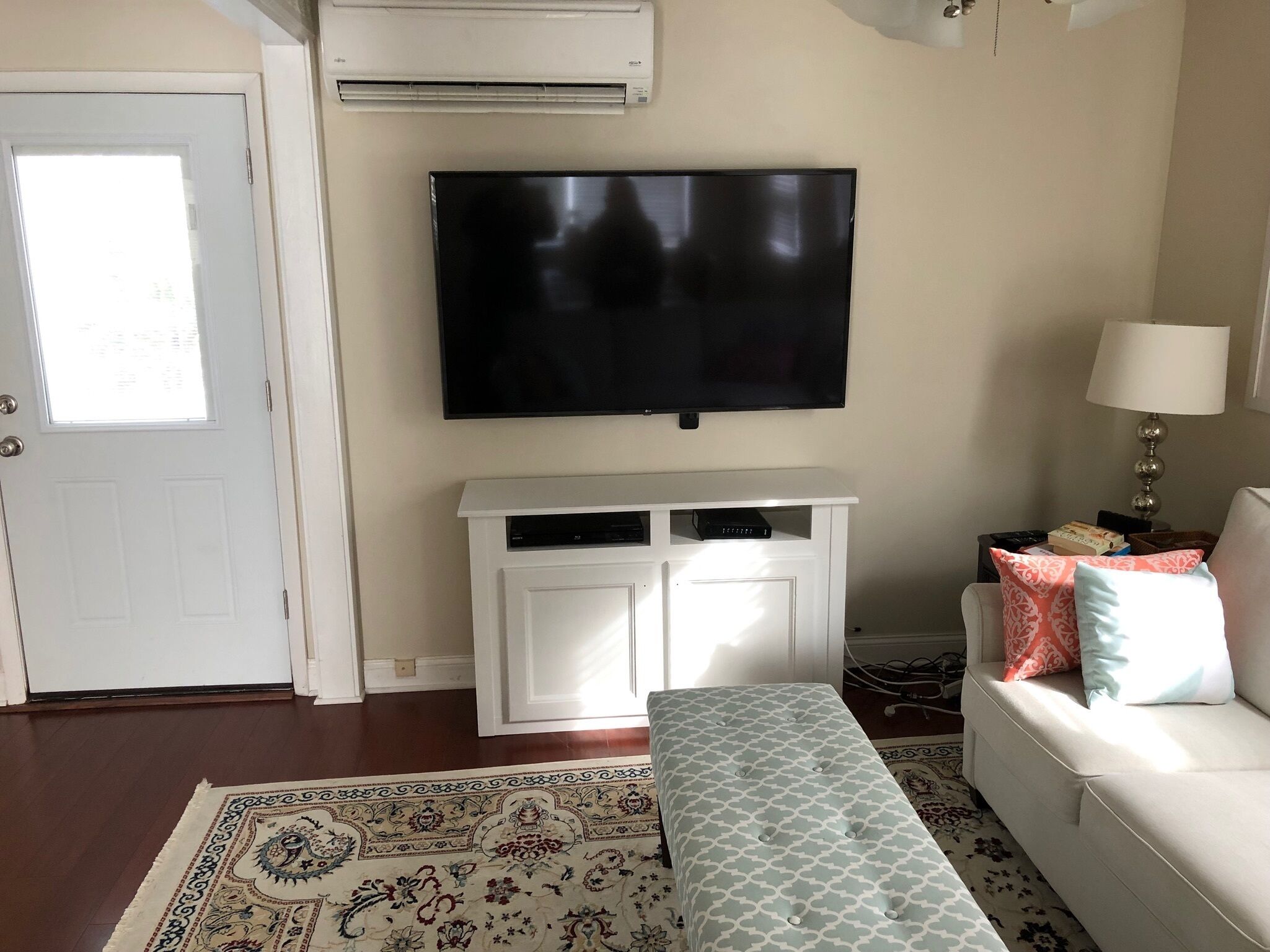 Ein Wohnzimmer mit einem wandmontierten Flachbildfernseher, einer weißen Unterhaltungseinheit darunter, einer Bank, einem gemusterten Teppich und einem weißen Sofa mit dekorativen Kissen.